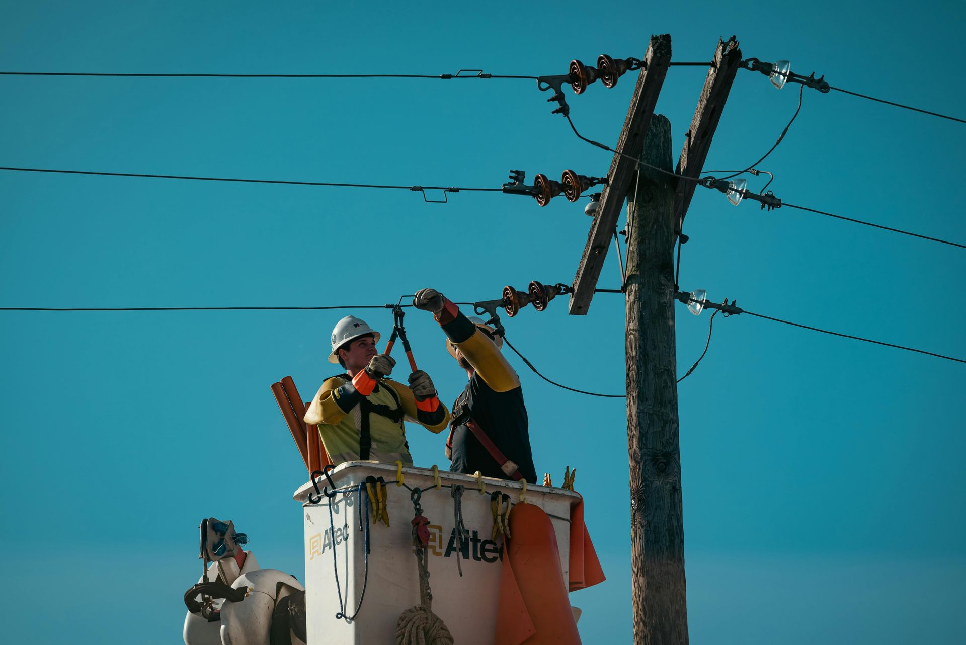 Utility workers repair power lines beside a pole, one in an orange vest, under a blue sky.
