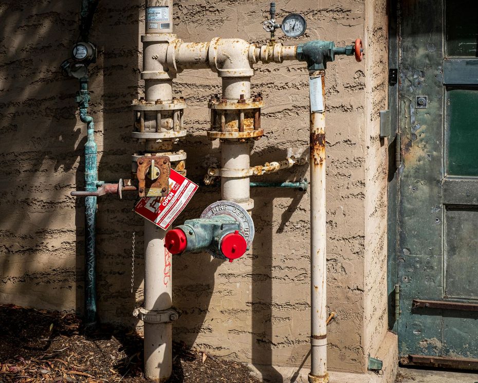 Rusty water pipes and valves mounted on a weathered wall, with a red meter and tags attached.