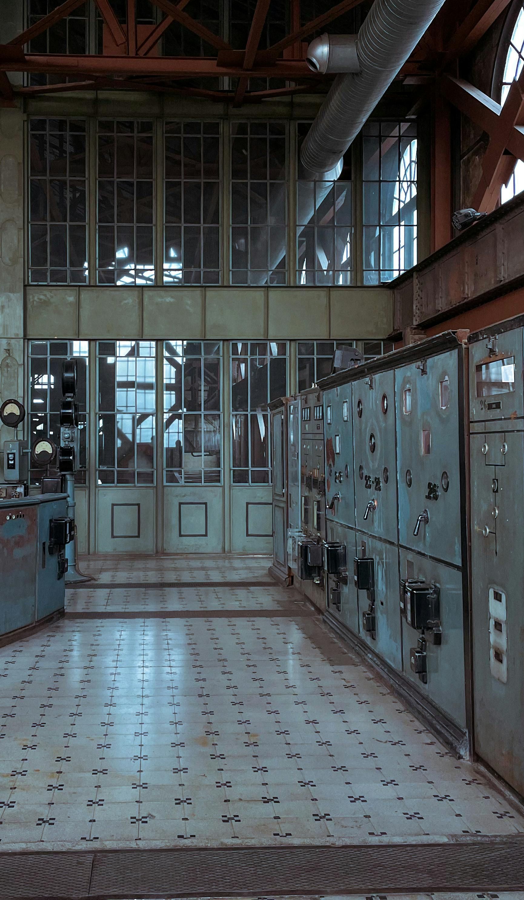Industrial control room with rows of electrical panels and a tiled floor under overhead pipes