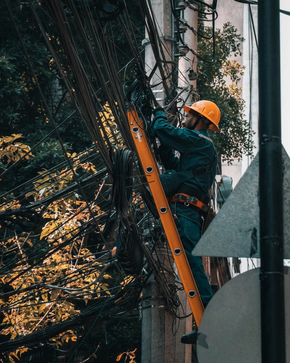 Worker in orange helmet climbs a ladder repairing cables on a utility pole outdoors.