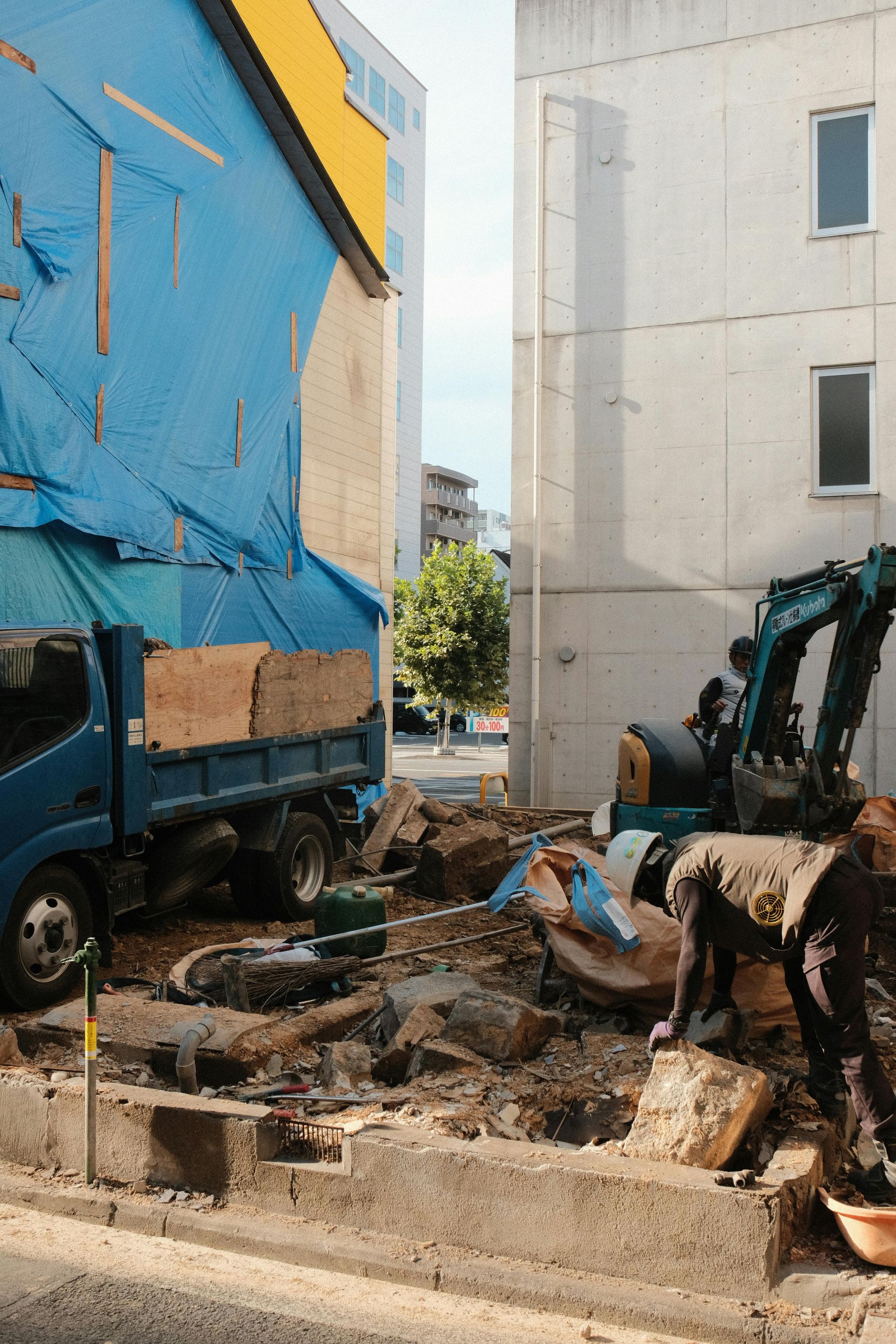 Construction site with excavator, dump truck, and debris between buildings
