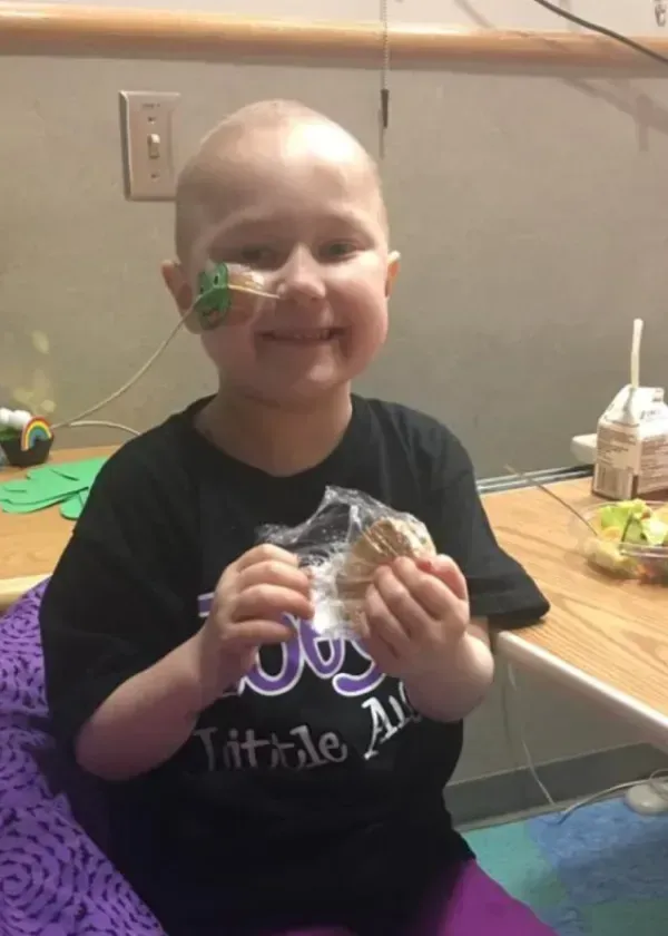 Child with bald head, holding food, smiles in hospital room; has tubes in nose.