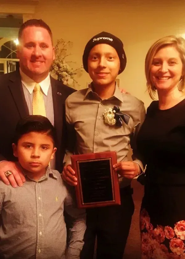 Family posing for photo; teen holding award, wearing beanie; adults smiling; young boy looking at camera.