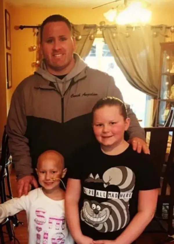 Man with two young girls, one with cancer, indoors, smiling.