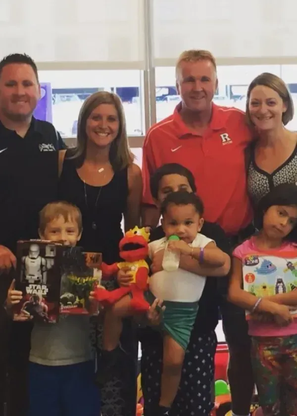 Group of adults and children smiling, holding toys, posing for a photo indoors, near a window.