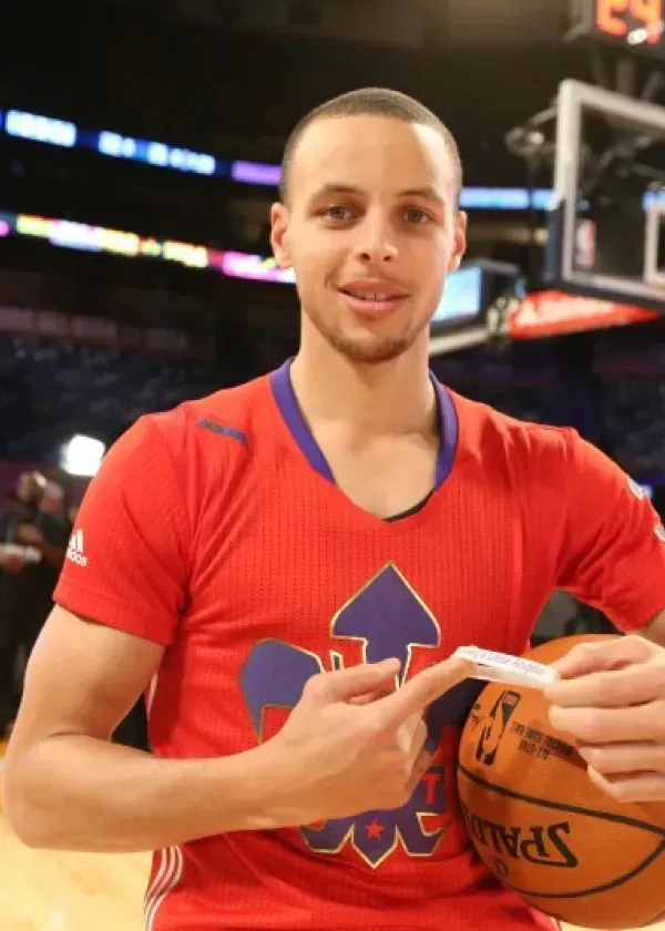 Stephen Curry holding a basketball, pointing to a piece of paper, smiling, on a basketball court.