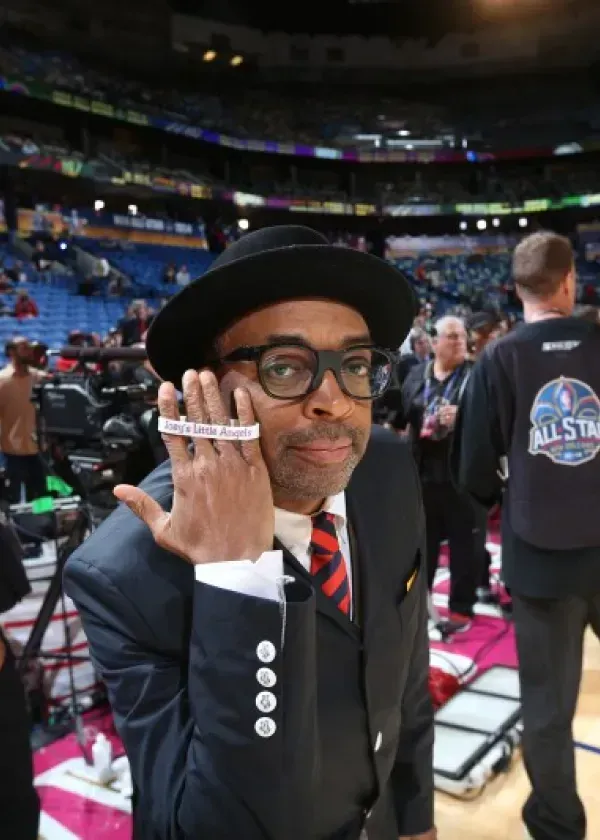 Spike Lee in suit, hat, and glasses, holding up a wristband at basketball game.