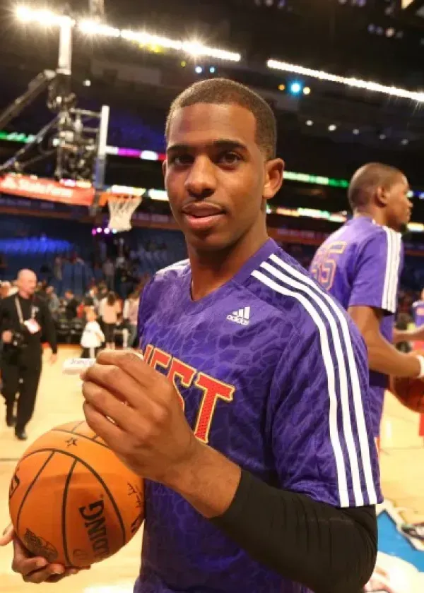 Chris Paul holding a basketball at an NBA All-Star Game. Purple Adidas jersey, hardwood court.