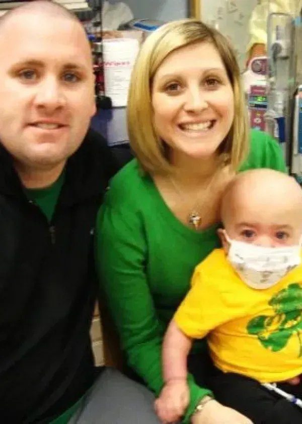 A family smiles: parents and child in hospital, child wearing a mask and yellow shirt.