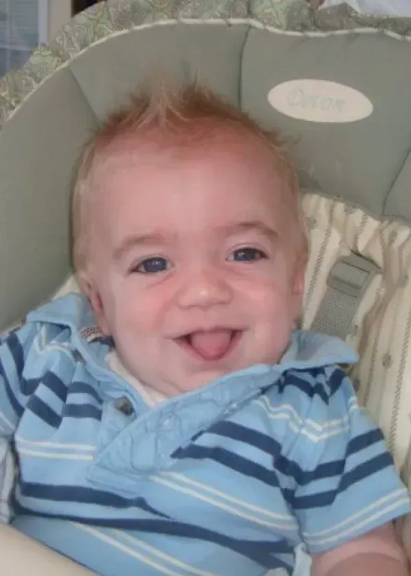 Smiling baby with blonde spiky hair, wearing a blue striped shirt, in a high chair.