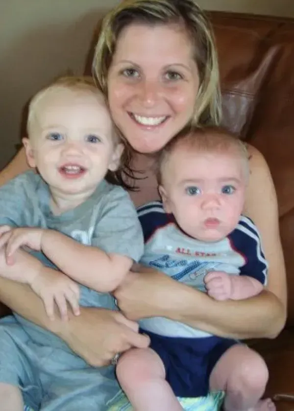 Woman smiling, holding two babies. One baby smiles, the other looks at the camera. All three are indoors.
