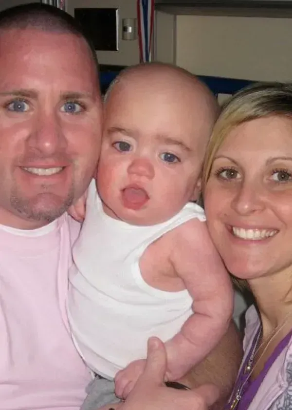 Man and woman smile, holding a baby wearing a white tank top. All three have light skin and are indoors.