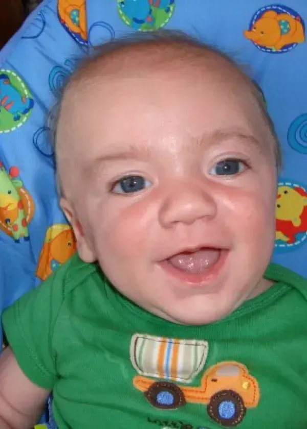 Smiling baby wearing a green shirt with a truck, in a blue and orange chair.