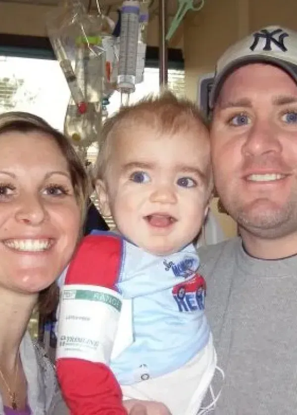 Family of three in a hospital room, smiling at camera. Young child being held by a parent. IV in the background.