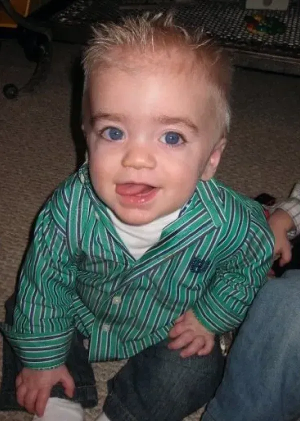 Blond-haired toddler in green striped shirt and jeans, sticking out tongue, looking up, indoors.
