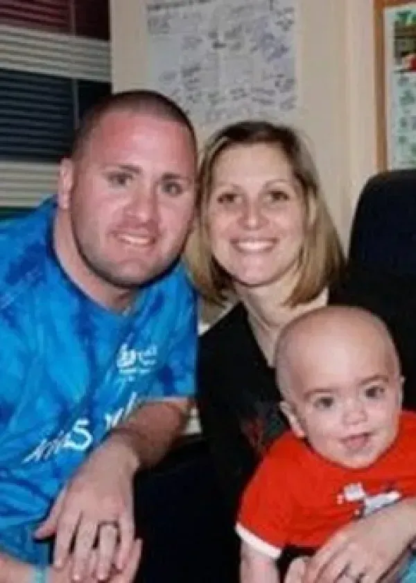 Family of three smiling, posing for a photo. Man in blue shirt, woman, and small child in a red shirt.