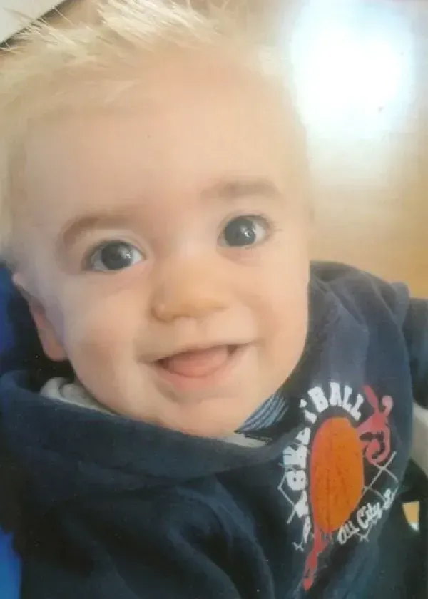 Smiling baby with blonde hair and blue eyes wearing a blue sweatshirt.