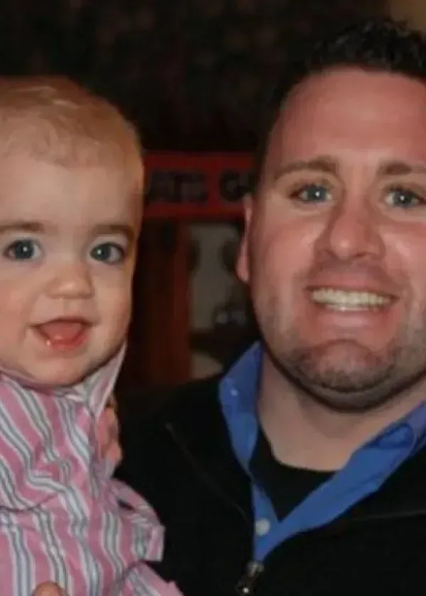 Man smiling with a toddler, both looking at the camera. The toddler is wearing a pink shirt.