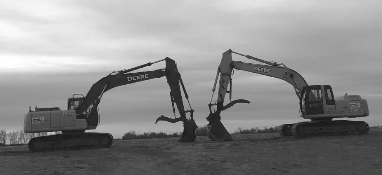 Two large construction excavators face each other in an open, overcast field, their buckets touching near the ground.