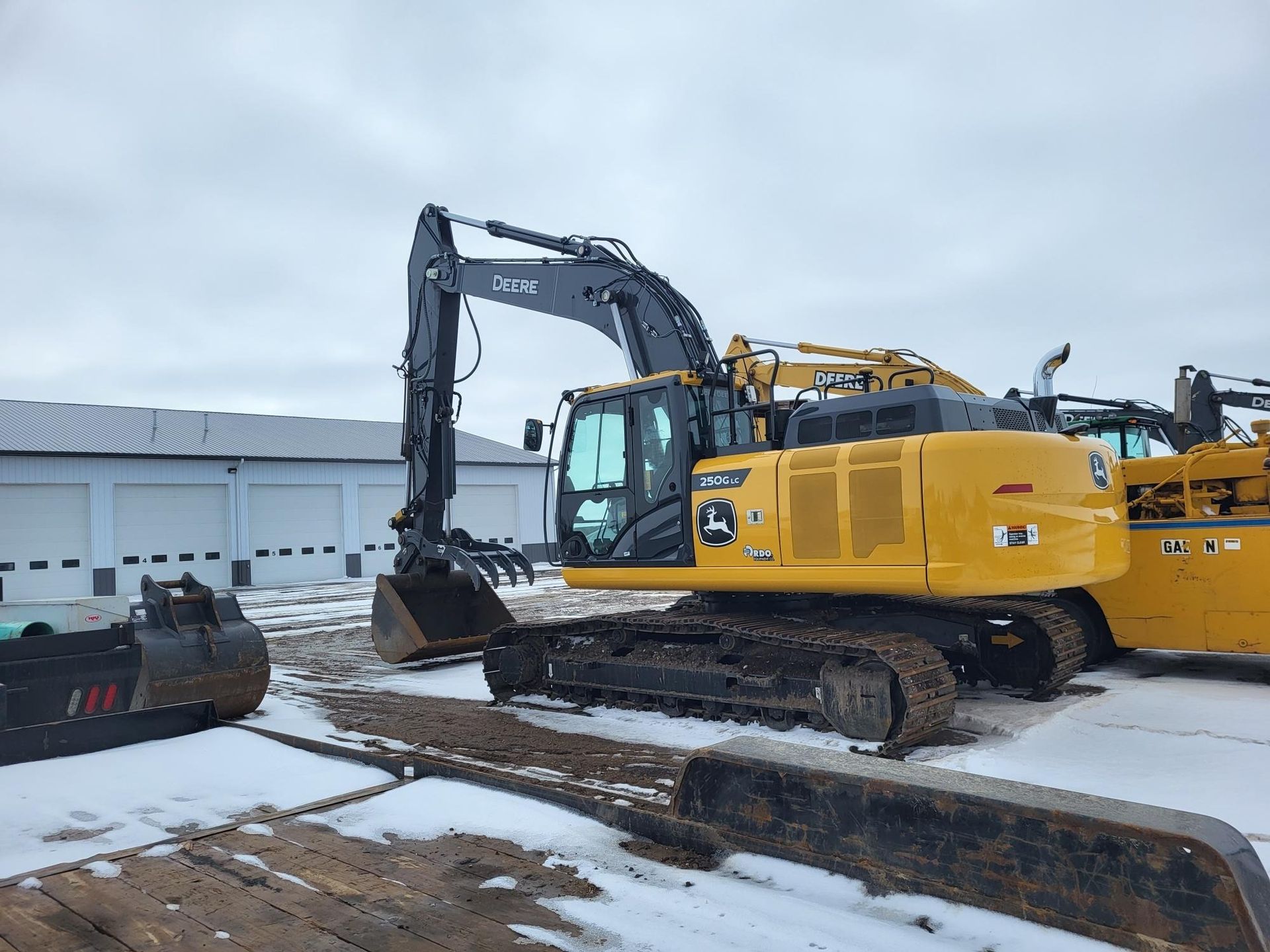 A yellow John Deere excavator parked on a snow-covered lot next to a building with multiple garage doors.