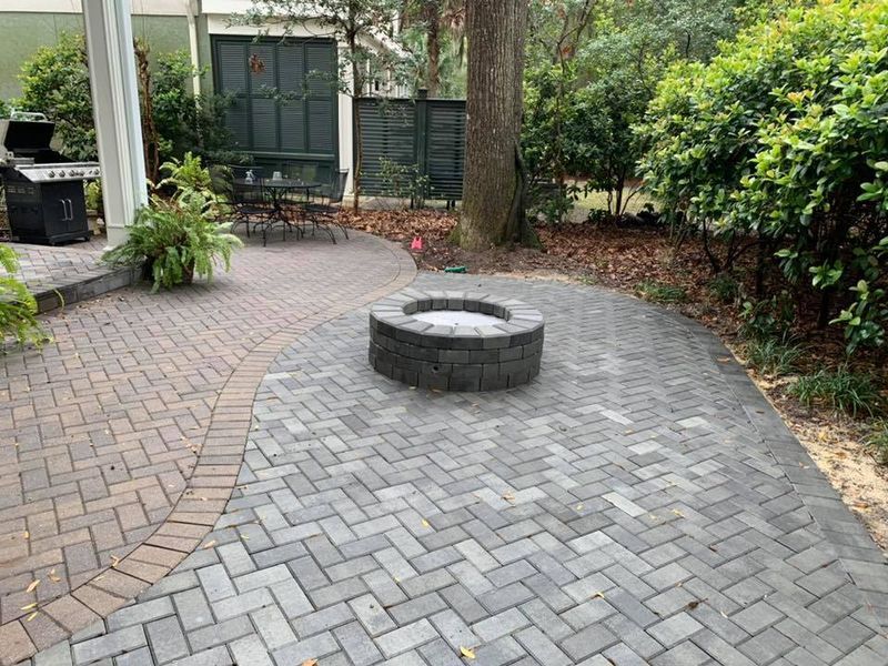 Brick patio with fire pit, grill, and outdoor seating surrounded by greenery.
