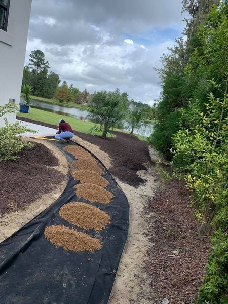A person kneeling, spreading mulch on a landscape bed beside a body of water.