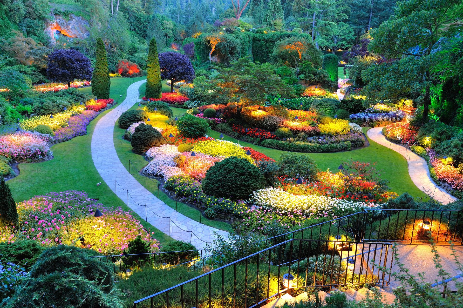 Pathway winds through a colorful, illuminated garden with lush flower beds and trees.
