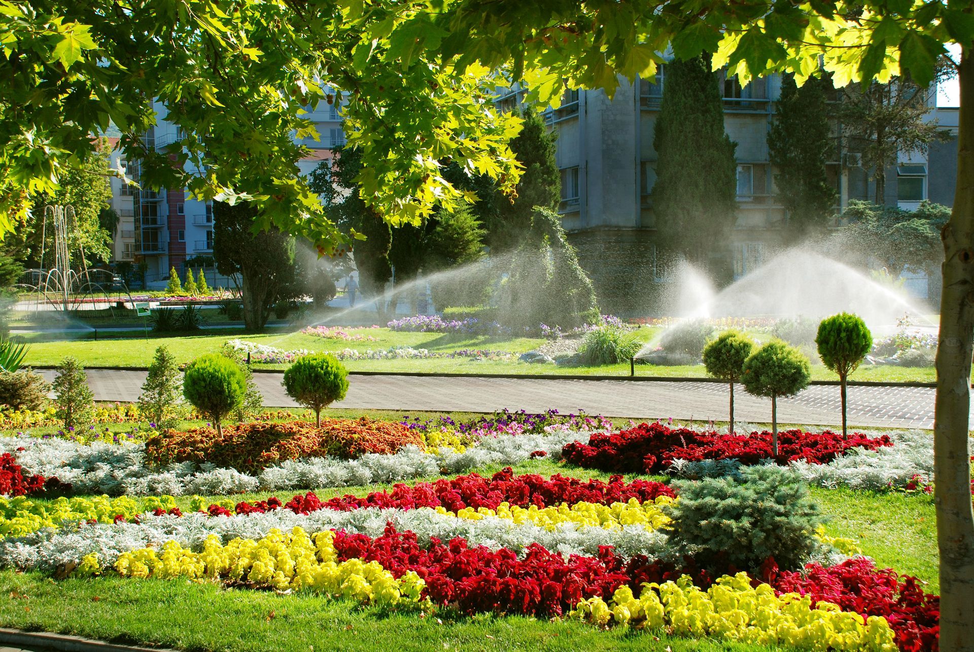 Lush flowerbeds in a park with sprinklers watering, under a canopy of green trees.