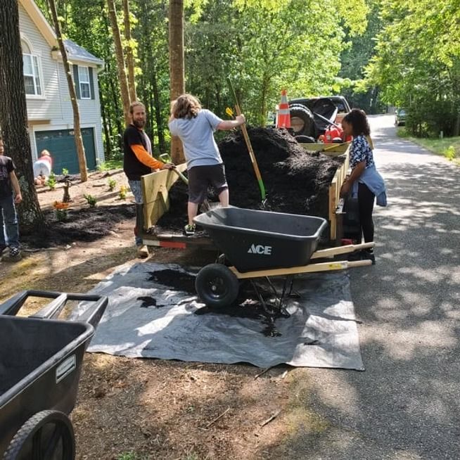 People loading a large black pile into a trailer with shovels on a tree-lined street