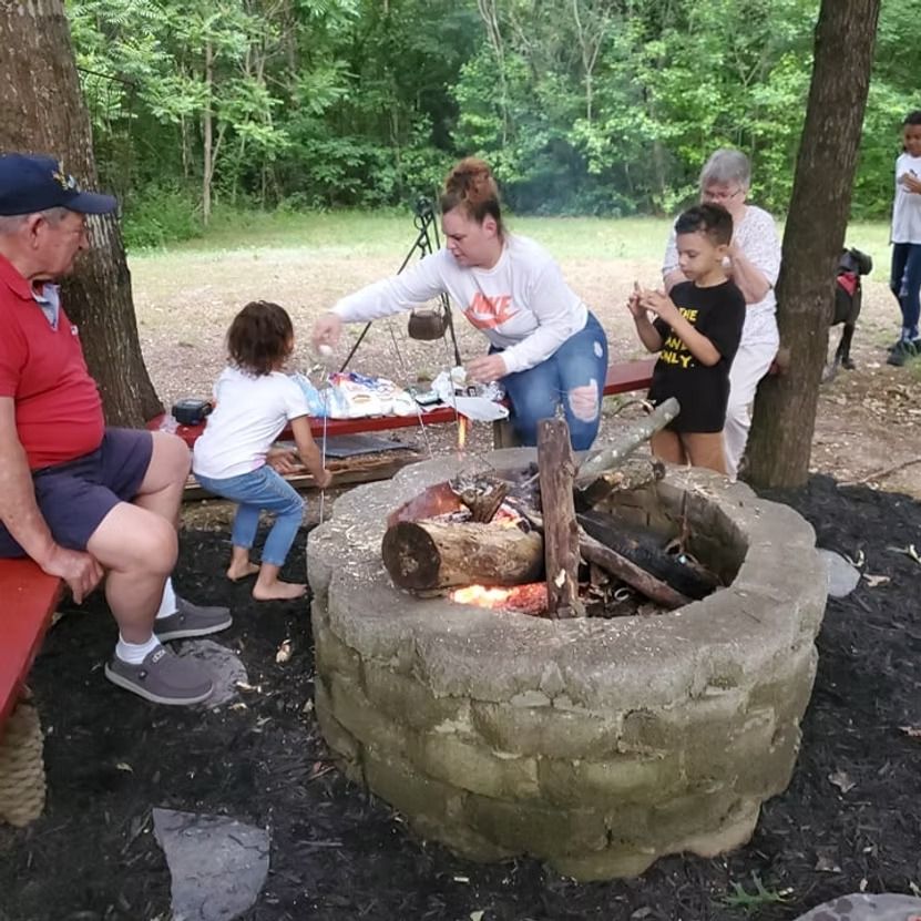 People gathered around a stone fire pit outdoors, roasting food at a picnic table in a wooded area.