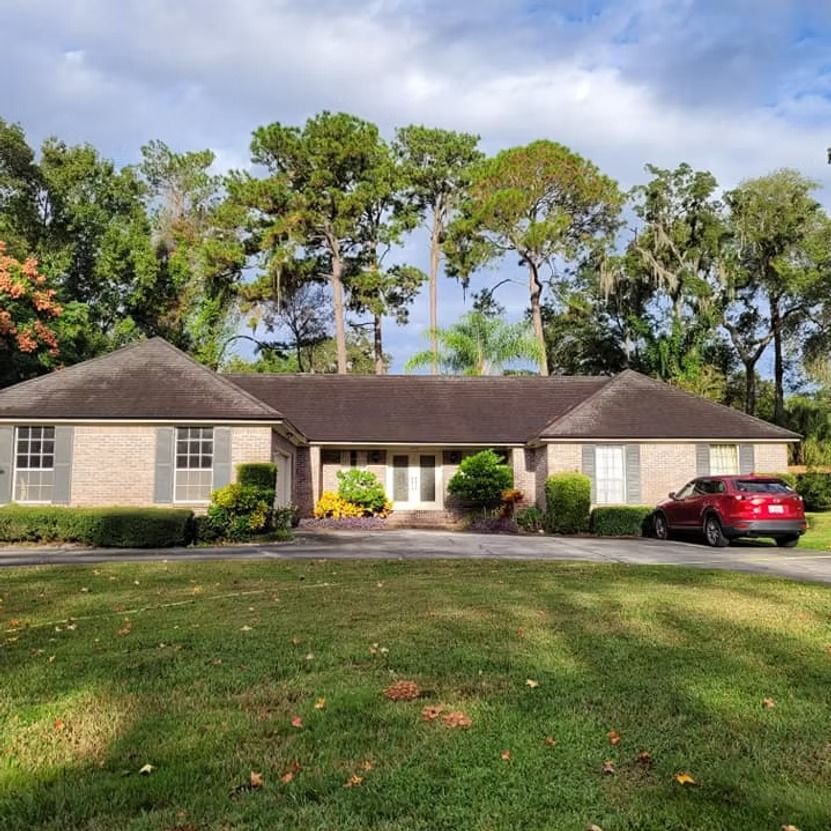 Single-story beige house with dark roof, trees behind, and a red car in the driveway.