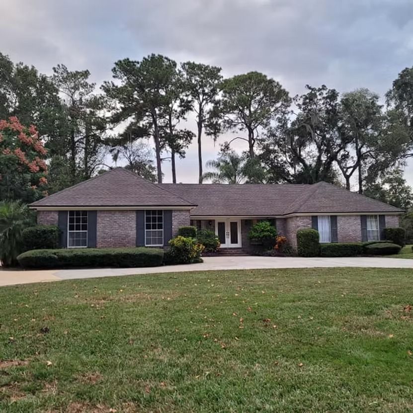 Single-story brick house with dark roof, front lawn, and tall trees in the background