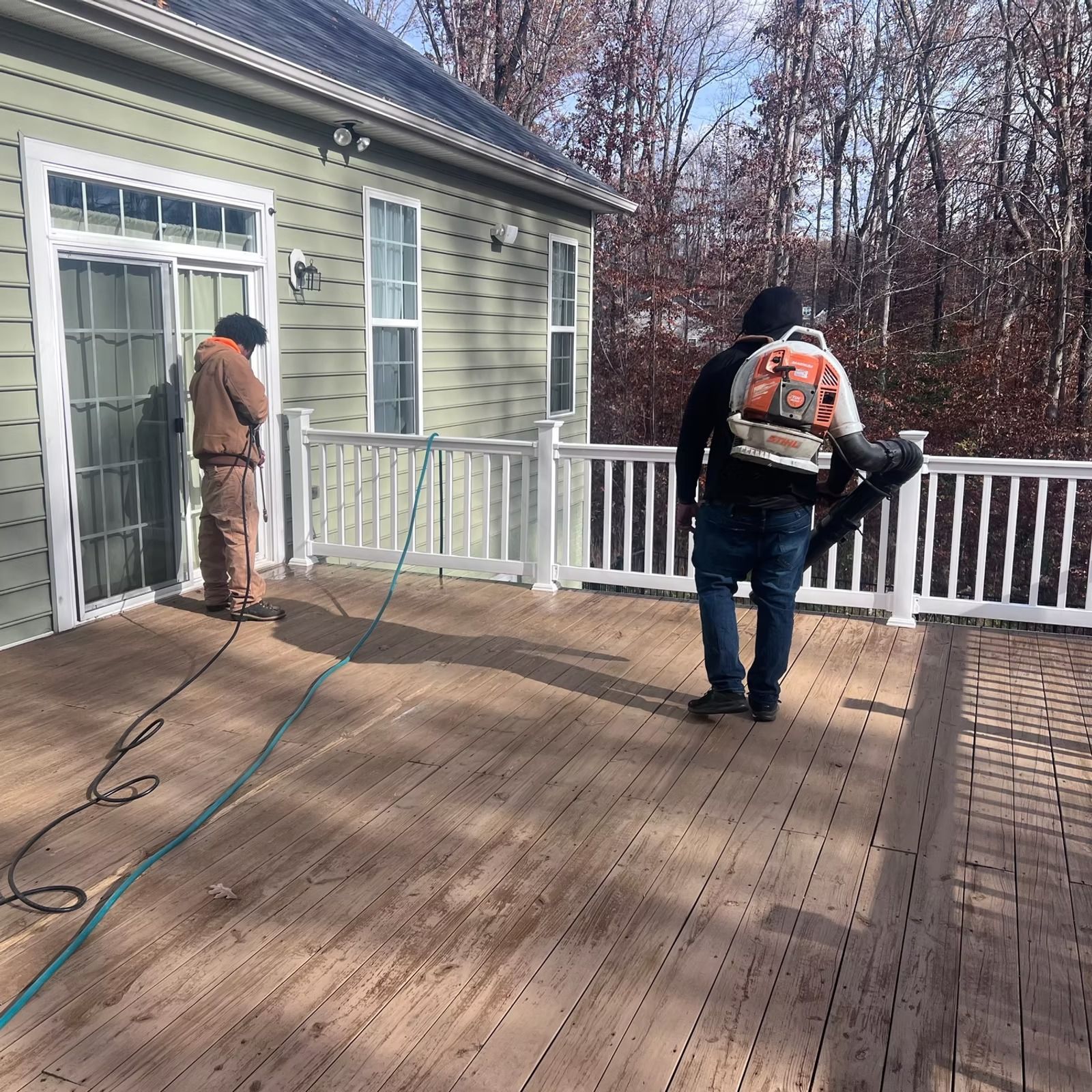 Two workers on a wooden deck beside a house, one carrying equipment and another near the door.