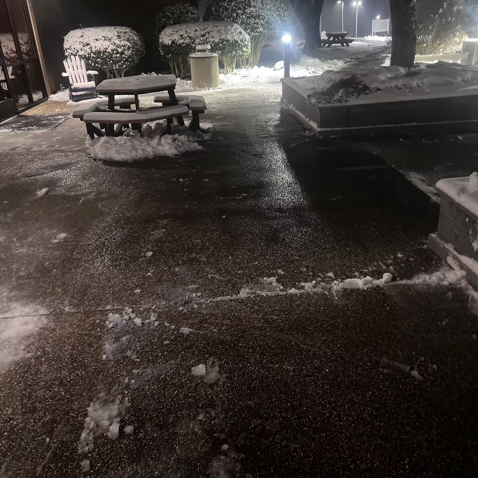 Snow-covered outdoor walkway with benches and planters at night, lit by bright lamps.