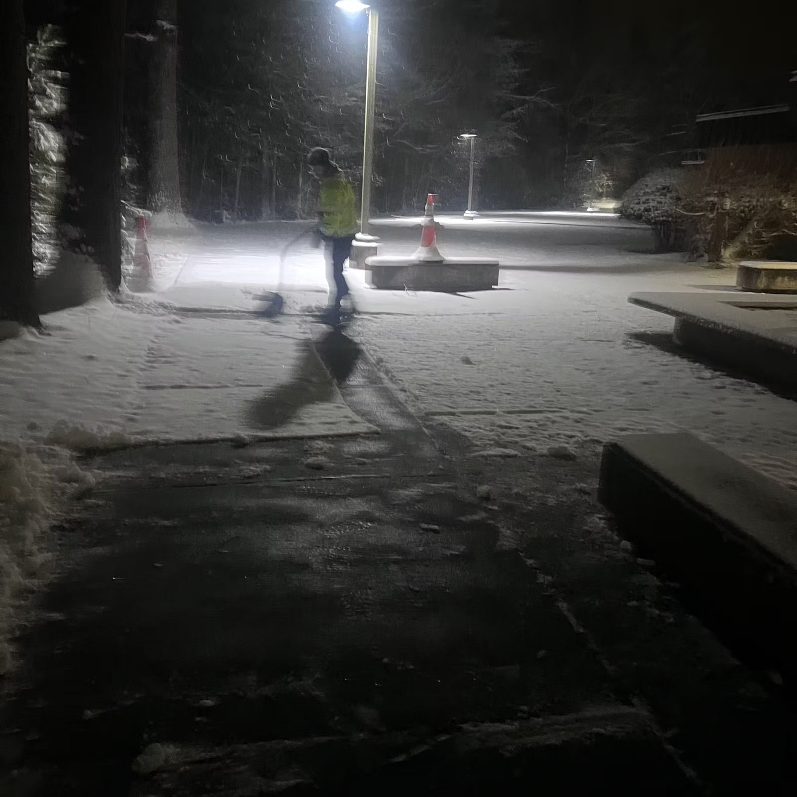 Snowy street at night with a person in a reflective jacket shoveling near a lamppost.