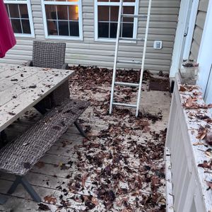 Leaf-covered patio with a white ladder, wicker table and bench beside a house exterior.
