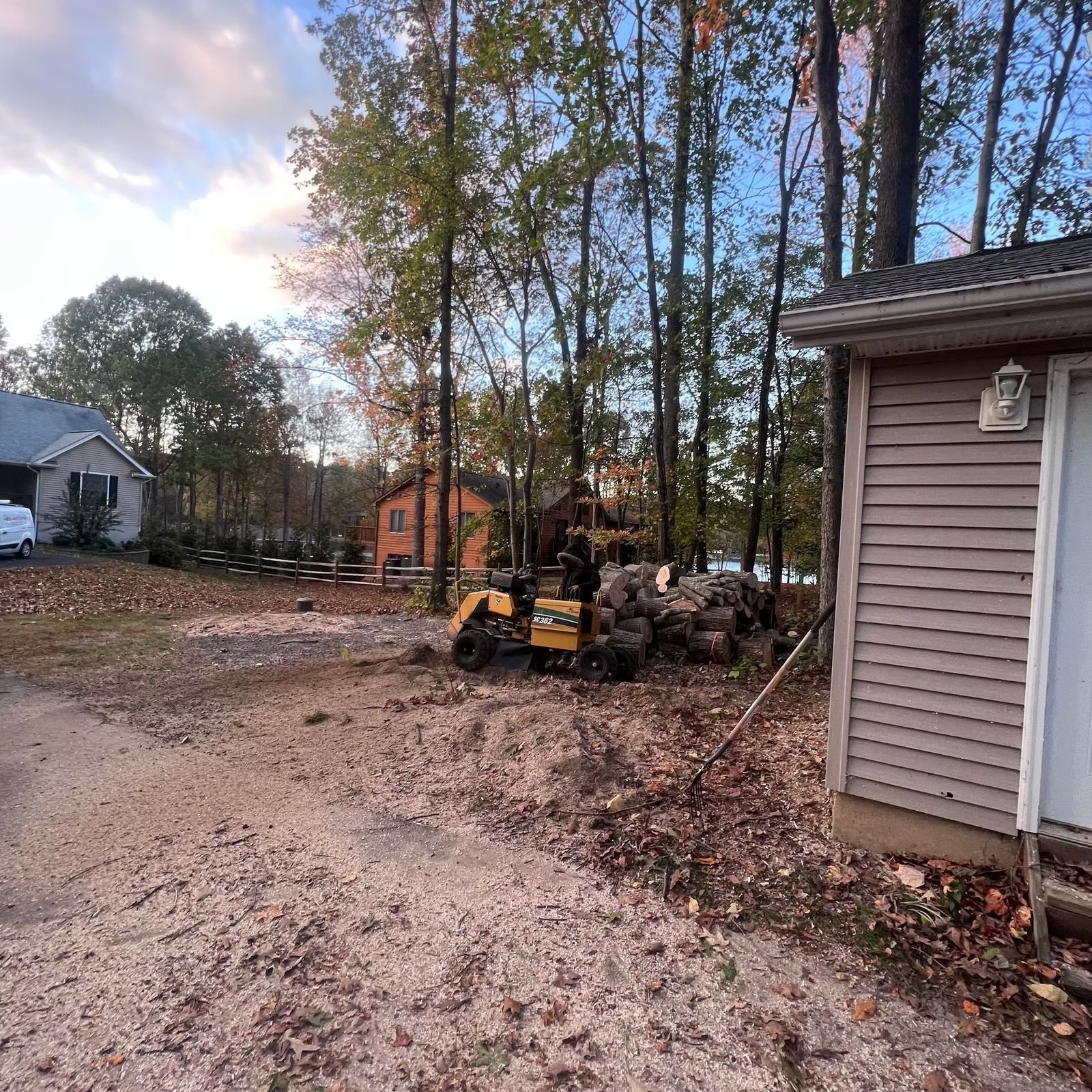 Gravel driveway beside a house, with a small yellow tractor and stacked logs in a wooded yard at dusk