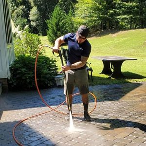 Person pressure-washes a stone patio with an orange hose on a sunny lawn.