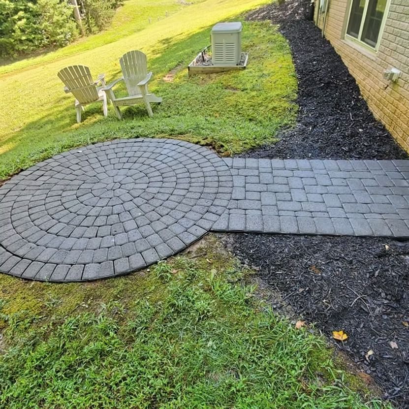 Circular stone patio and paver walkway beside a brick house, with lawn chairs and an outdoor AC unit.