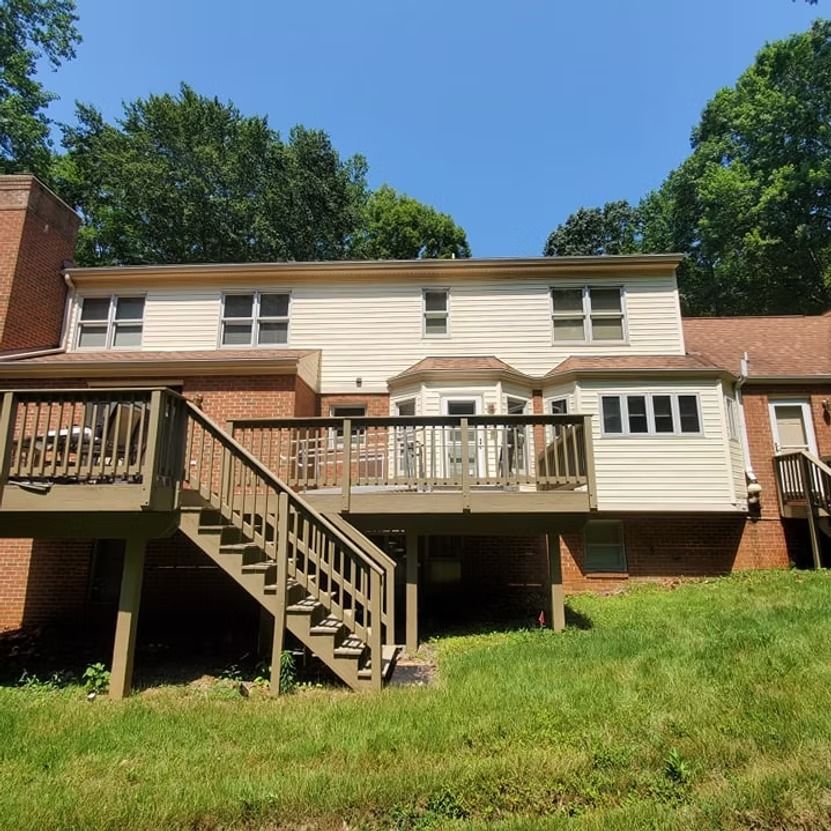 Rear view of a house with wooden deck stairs and railings, set on a grassy slope under a blue sky
