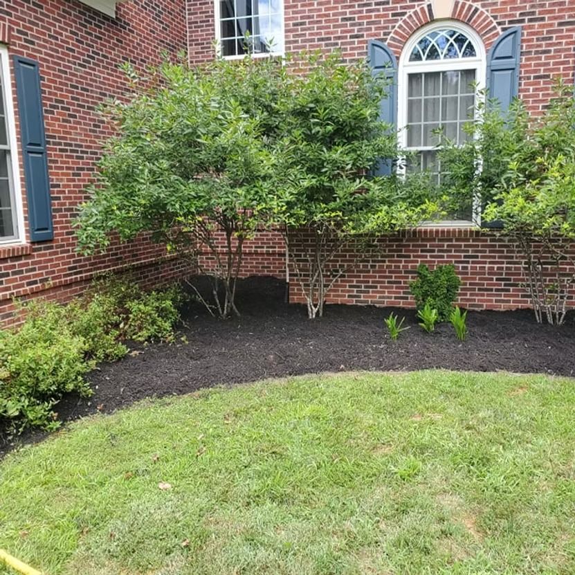 Brick house garden bed with shrubs, dark mulch, and green lawn beneath an arched window