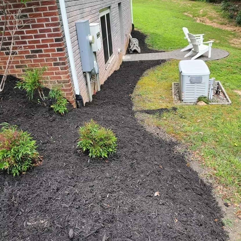 Mulched garden bed along a brick house with small shrubs and a walkway to patio chairs