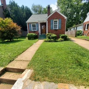 Small red brick house with blue shutters, front lawn, and concrete walkway leading to the porch