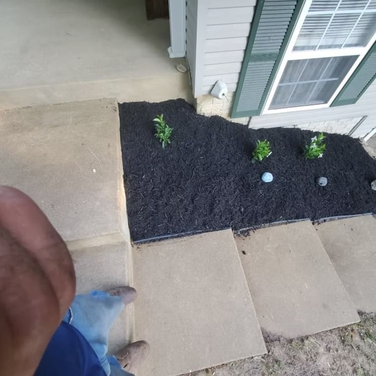 Front porch with tan pavers and black mulch bed, small green plants, and a person’s hand and blue pants at left.