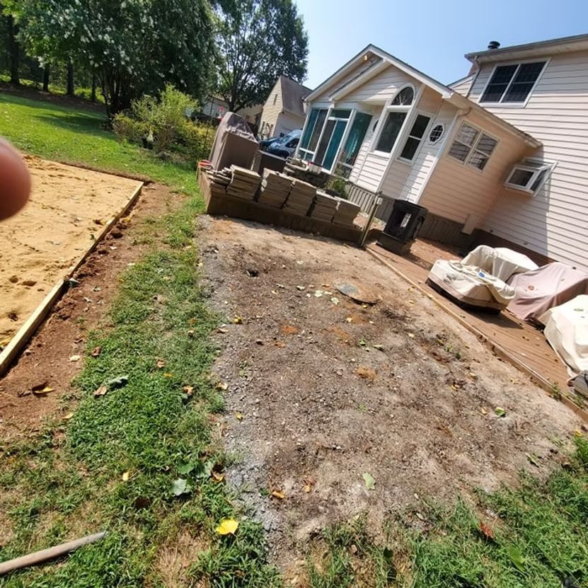 Backyard with a patio, bare dirt area, and stacked materials beside a white house