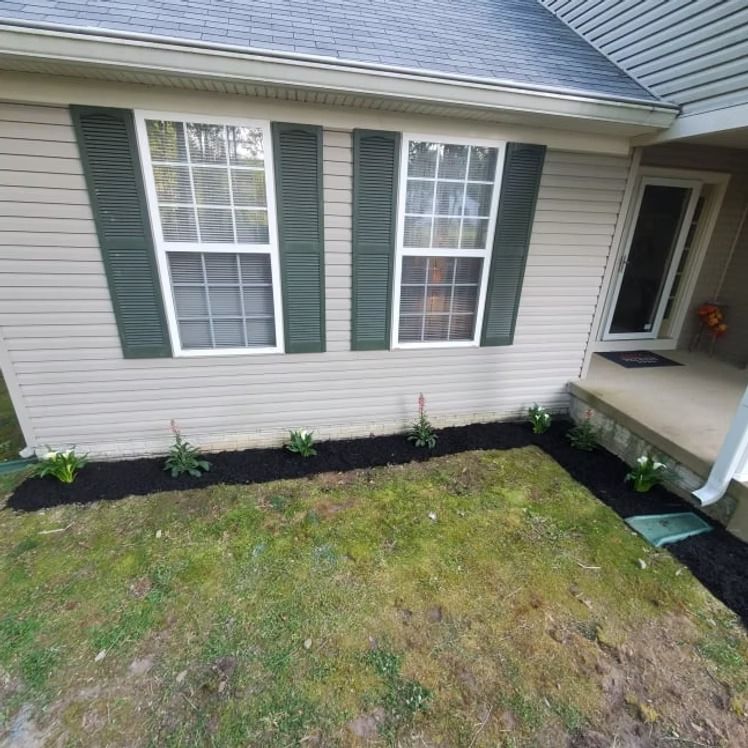 House front with two windows, fresh black mulch border, small plants, and a concrete porch beside a patchy lawn.