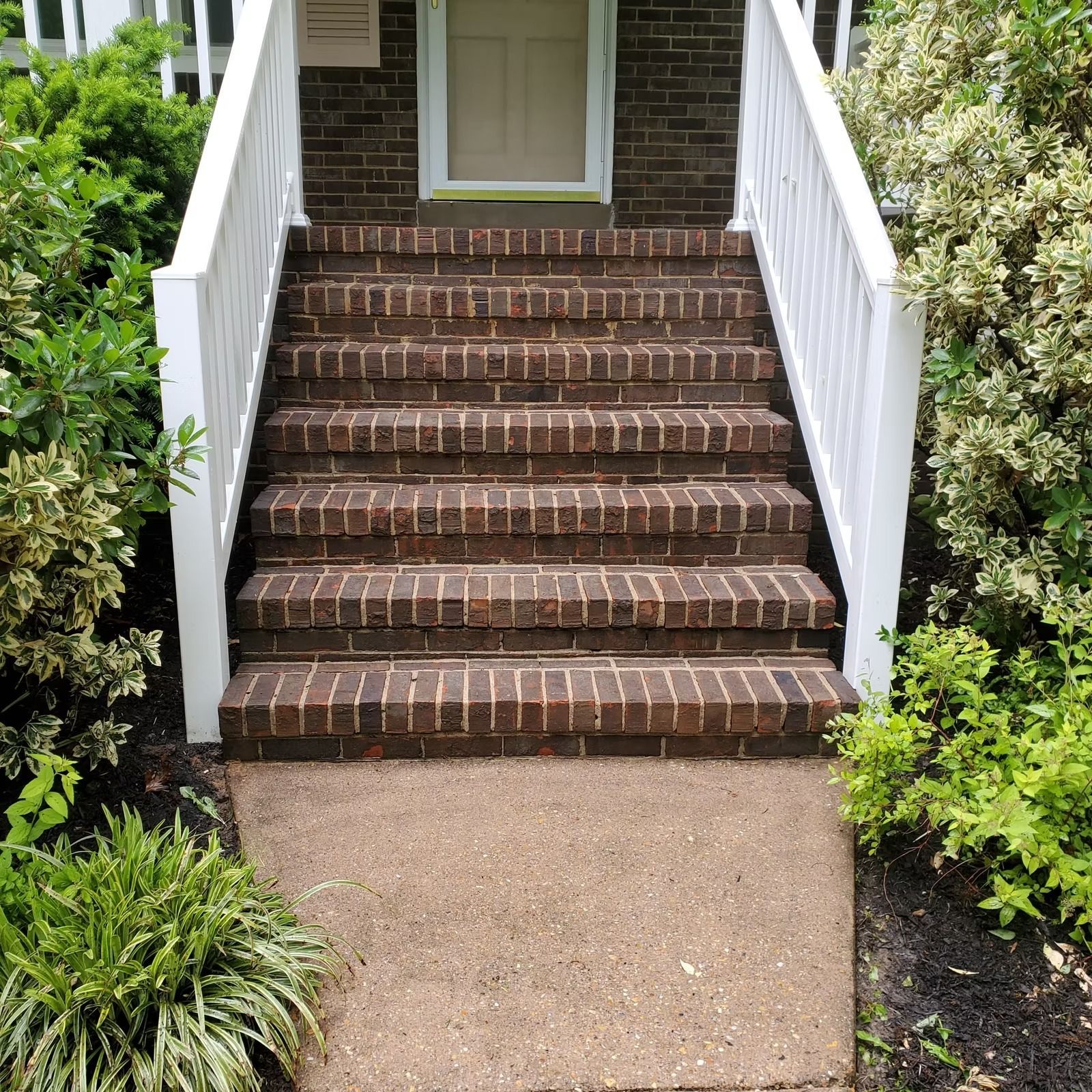 Brick steps with white railings leading up to a front door, surrounded by greenery.
