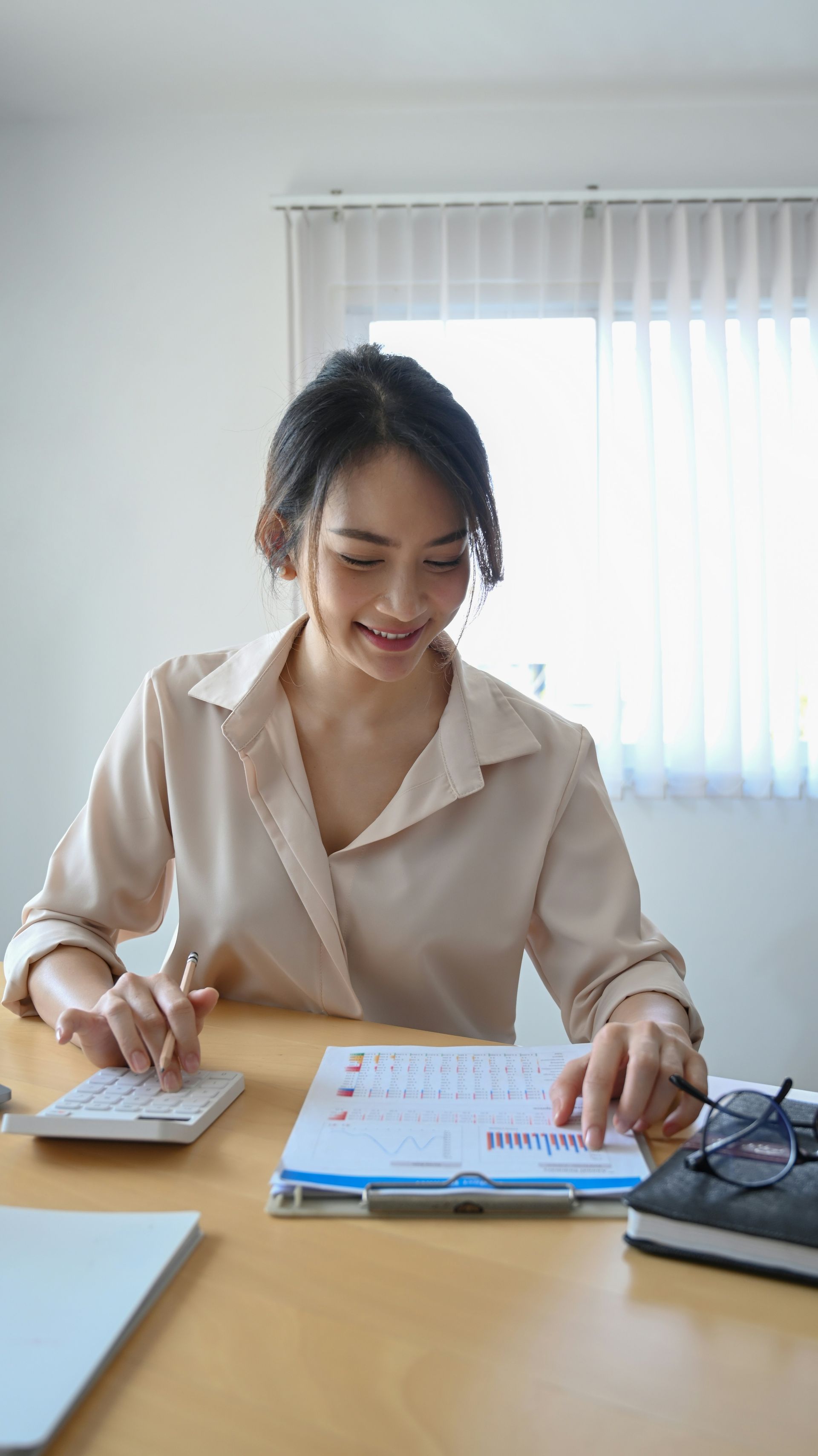 Woman writing on paper at a desk with a calculator and laptop in a bright office