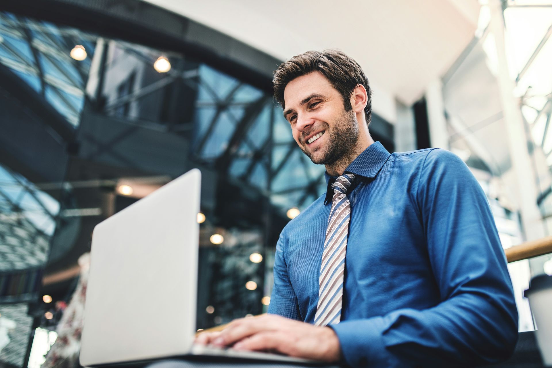 Man in a blue shirt smiling while using a laptop in a modern office lobby