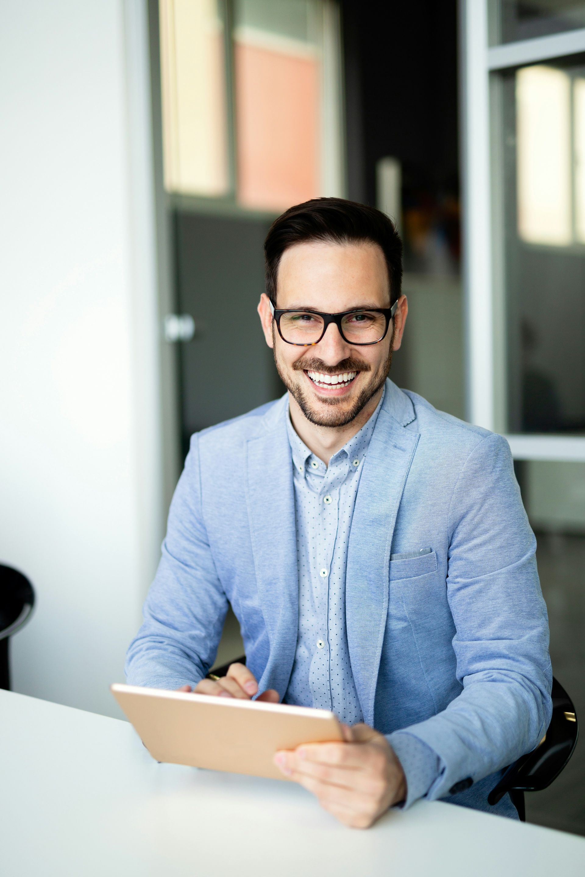 Smiling man in glasses holding a tablet at a desk in a bright office